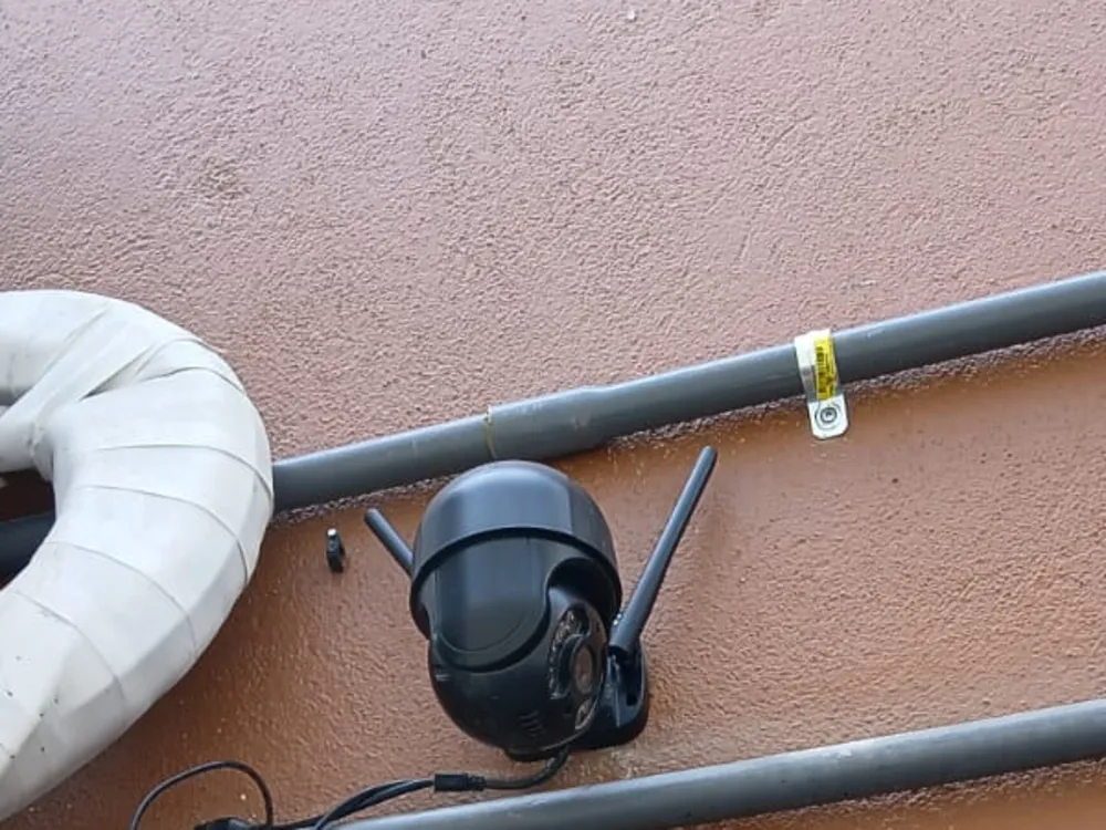 Cámara PTZ WiFi negra con dos antenas montada en un muro de estuco junto a la fuente de poder, entre dos tubos